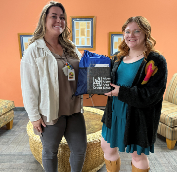 Two white women with an orange wall holding a bag with AAACU logo. Women on the left is a blonde and taller than women on the right who is readhead. 